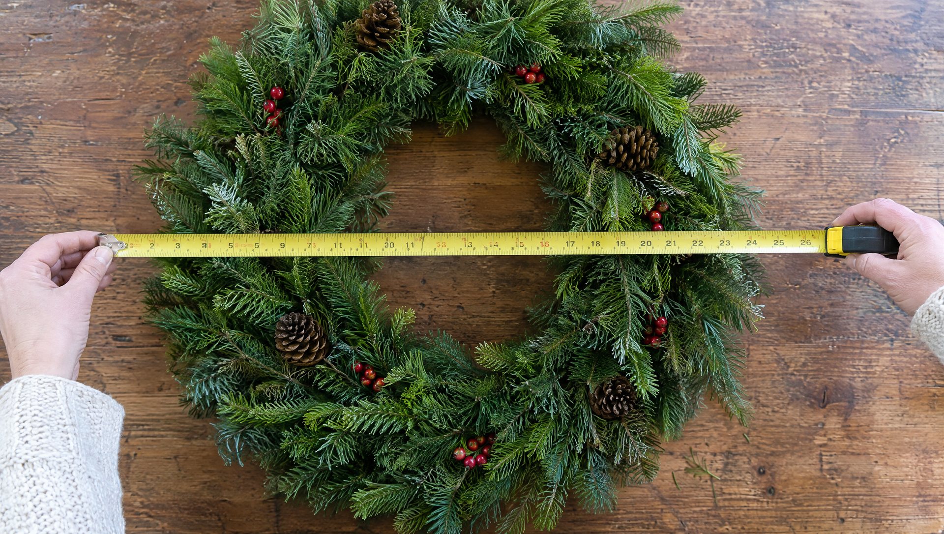 Hands measuring a Christmas wreath across its foliage with a tape measure