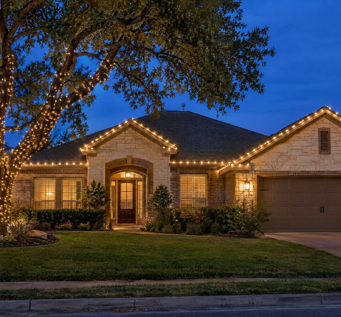 One-story Austin limestone and brick home at blue hour with warm white roofline lights and a wrapped live oak