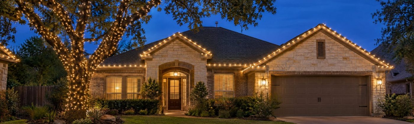 One-story Austin limestone and brick home at blue hour with warm white roofline lights and a wrapped live oak