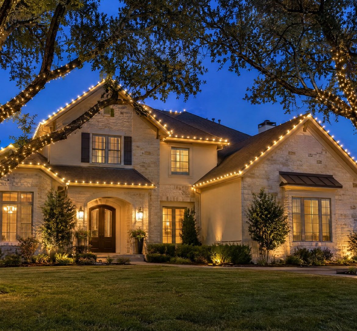 Two-story Austin home at blue hour with lit rooflines and two live oaks wrapped in warm white lights.