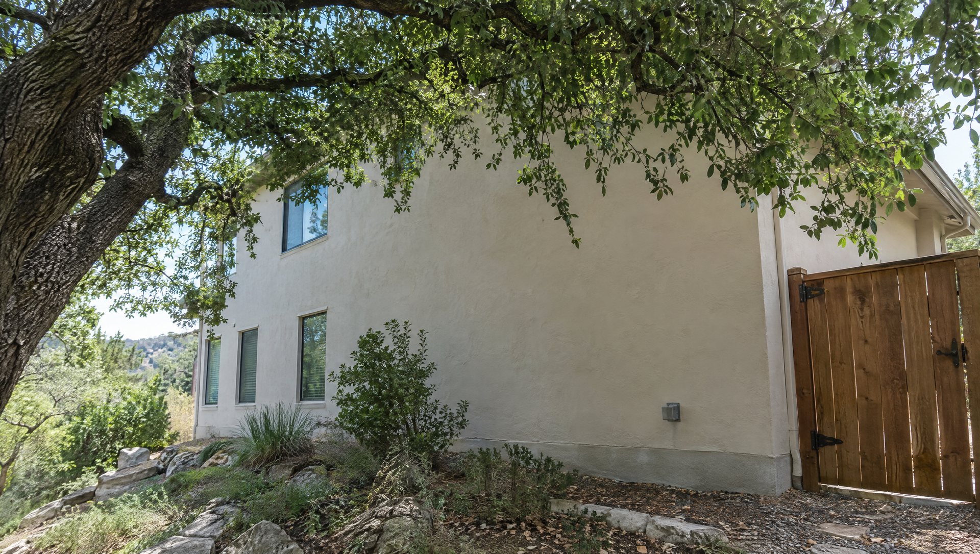 Side of a stucco house on a slope with an outdoor outlet and a live oak canopy hiding the roof edge.