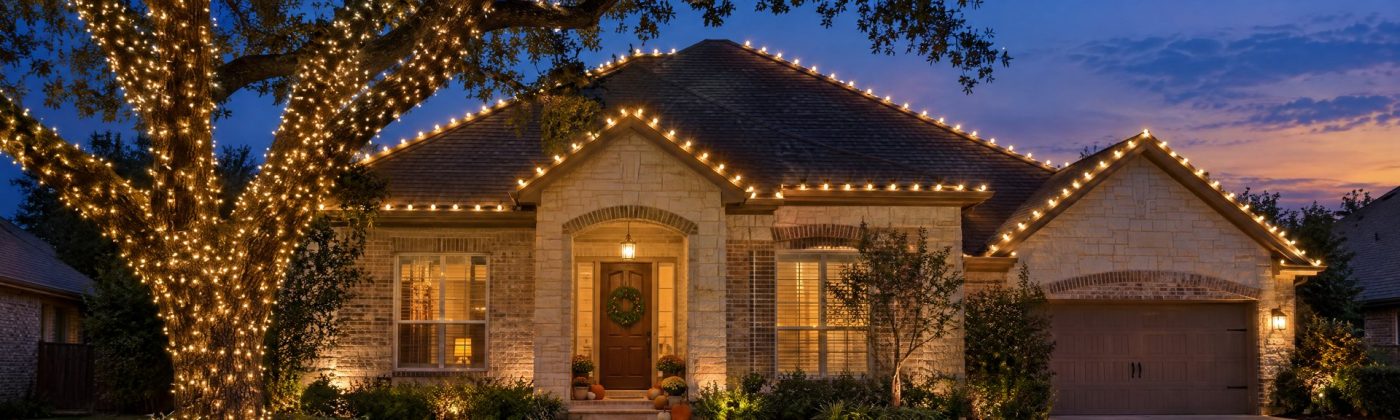 A limestone home at blue hour with warm white roofline lights, a wrapped live oak and pumpkins on the porch steps before Thanksgiving.