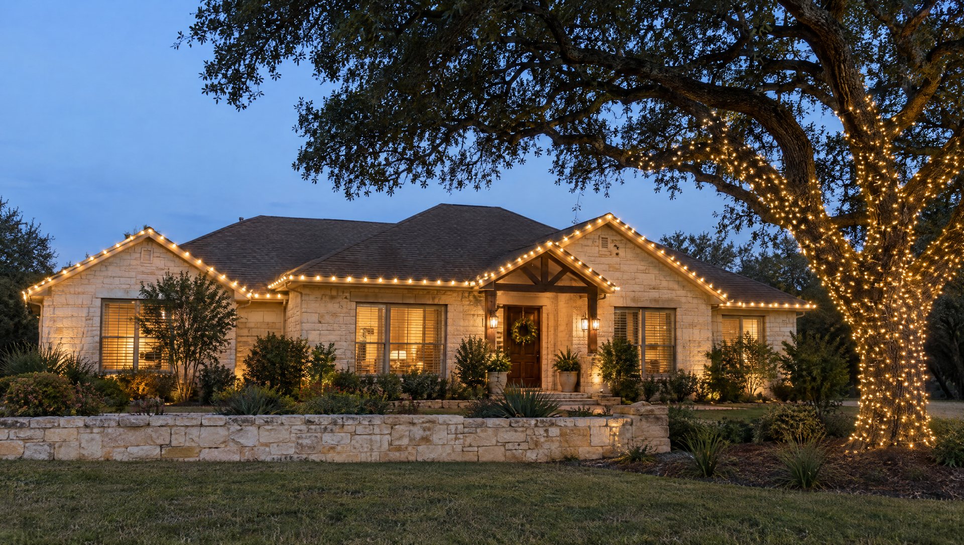 A one-story limestone house at dusk with warm white roofline bulbs, a wreath on the door and a wrapped live oak trunk.
