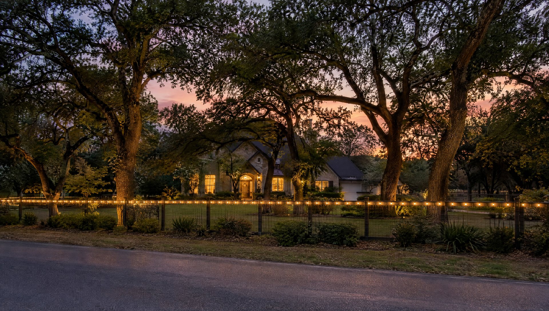 Fence with Christmas lights along the street in front of a home set back behind live oaks at dusk