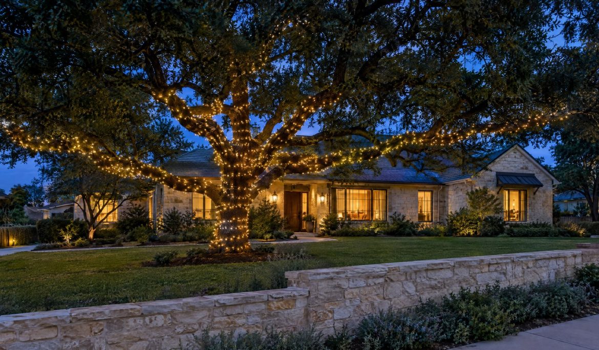 Live oak at blue hour with its trunk and long limbs wrapped in warm white lights and the crown left dark.