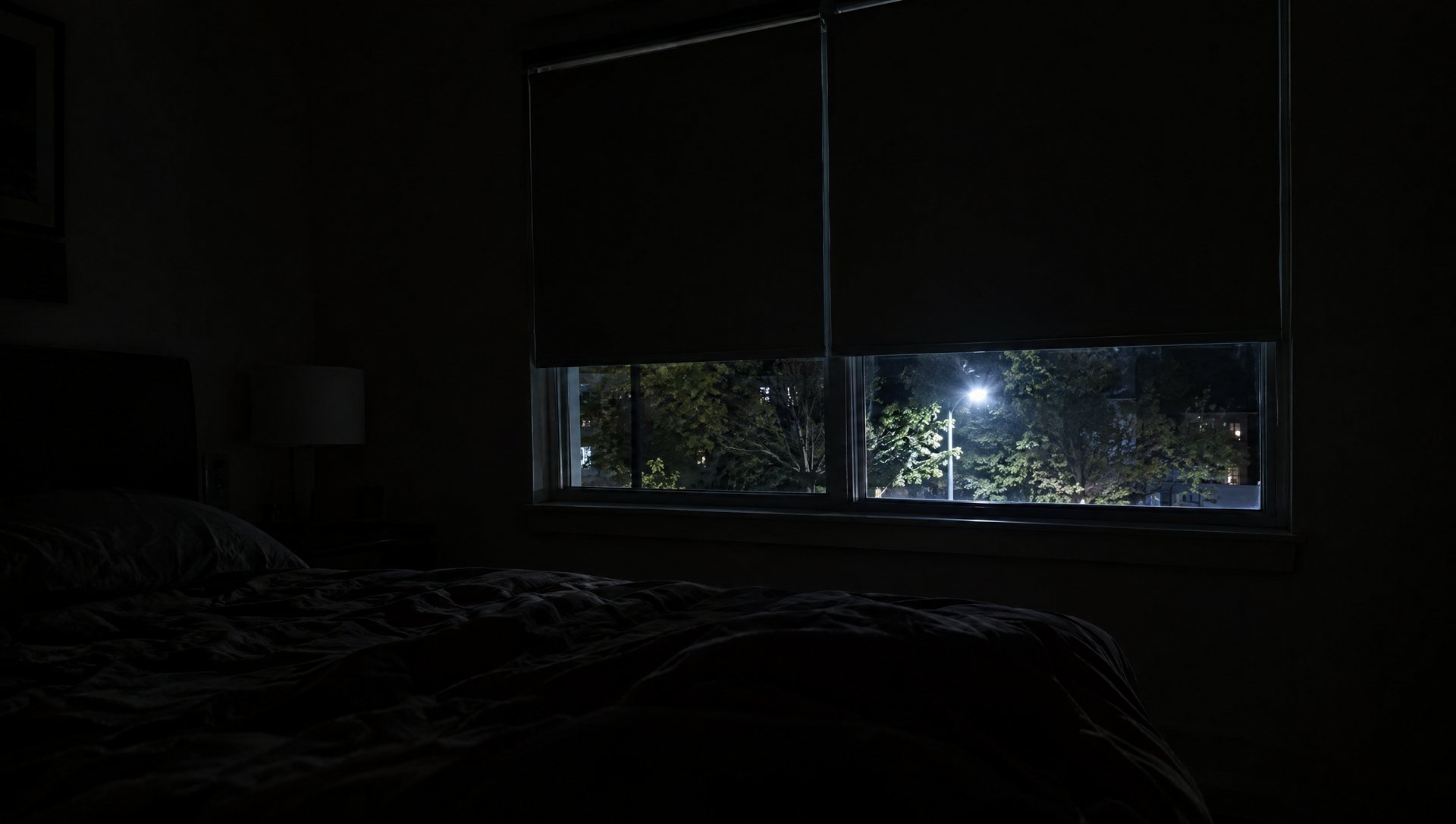 Dark bedroom with blackout shades partly drawn and a streetlight glowing through the gap