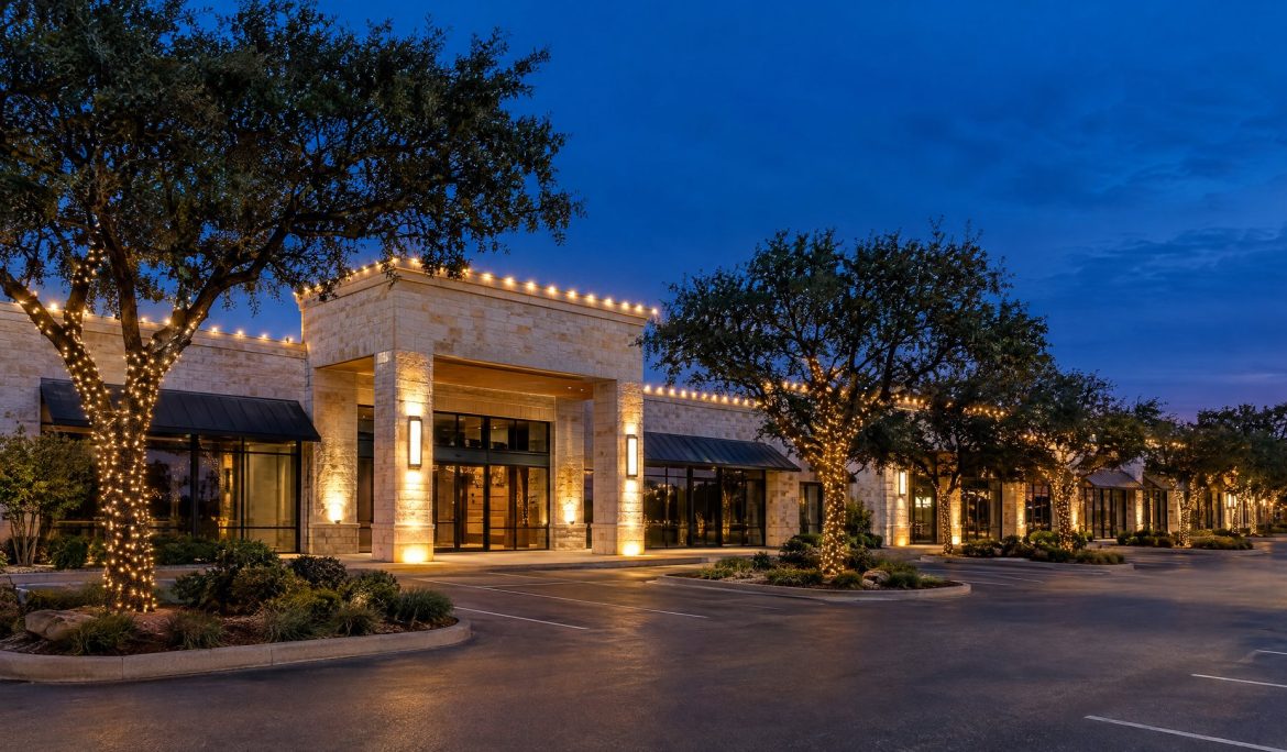 Limestone retail center at blue hour with warm white parapet lights and wrapped parking-island live oaks.