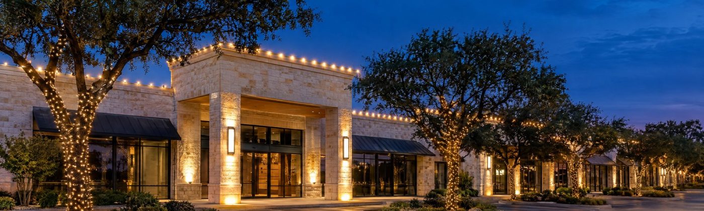 Limestone retail center at blue hour with warm white parapet lights and wrapped parking-island live oaks.