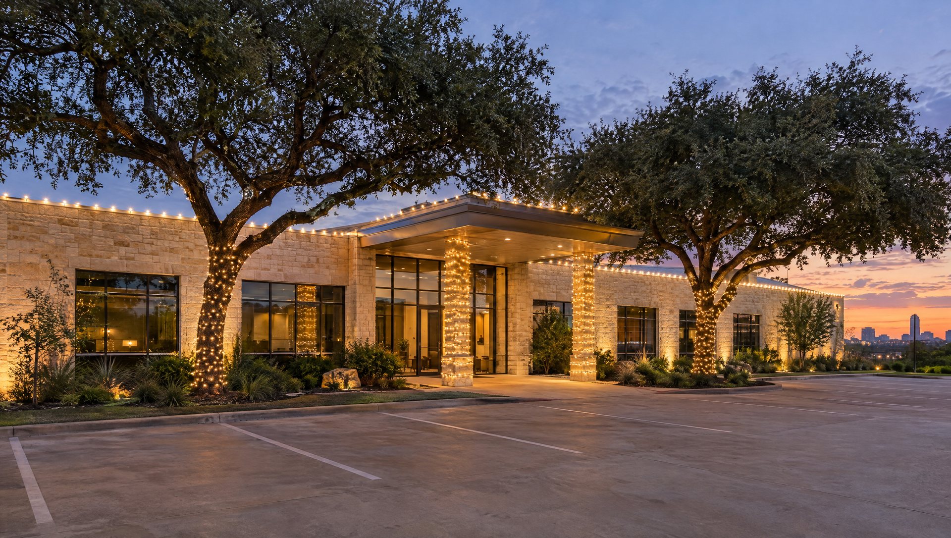 A limestone and glass office building at dusk with lit roofline, wrapped entry columns and two wrapped live oaks.