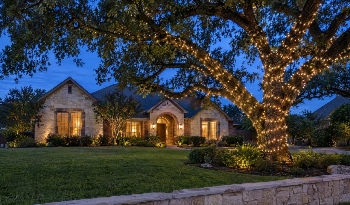 Large live oak at blue hour with its trunk and main limbs wrapped in warm white lights in front of a limestone home.