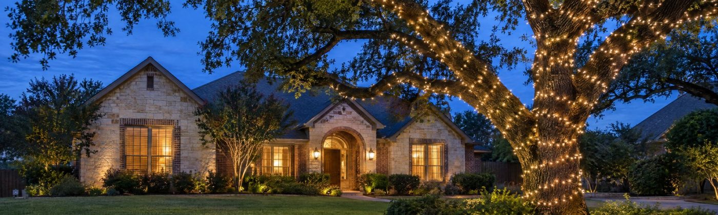 Large live oak at blue hour with its trunk and main limbs wrapped in warm white lights in front of a limestone home.