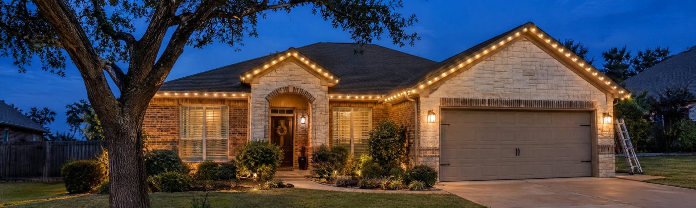 One-story brick and limestone home at blue hour with warm white C9 lights along the roofline and gables.