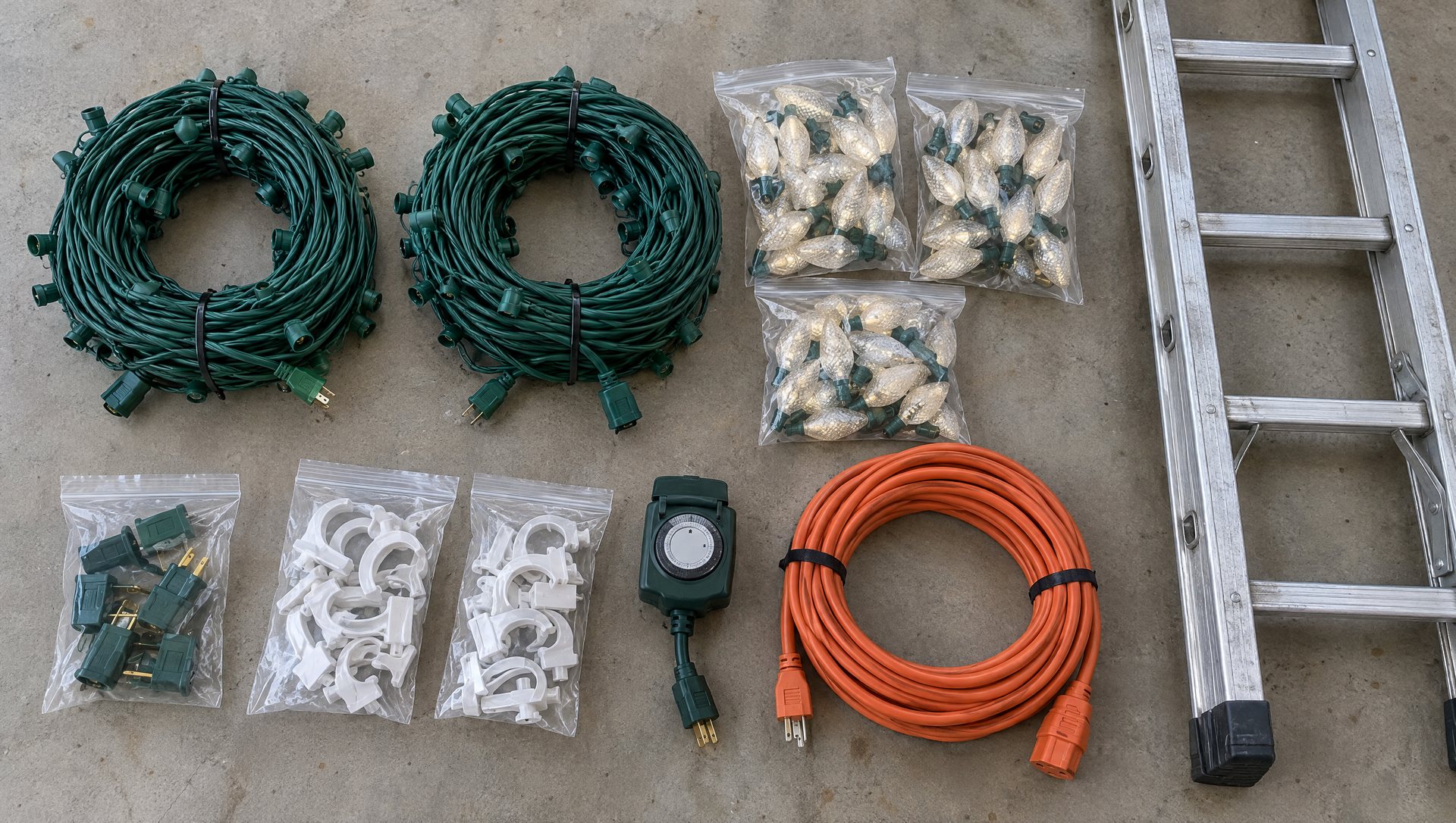 DIY Christmas light materials on a garage floor: coiled C9 cords, bags of bulbs and clips, plugs, a timer and a ladder.