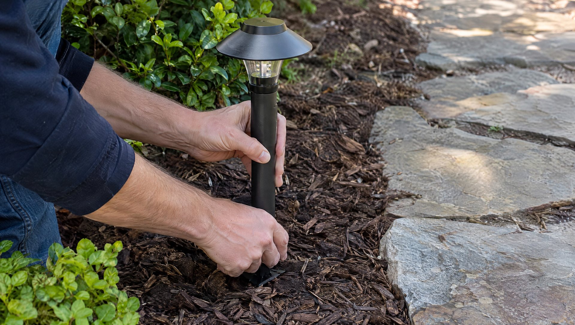 Hands pressing a path light stake into a mulched bed beside a stone walkway