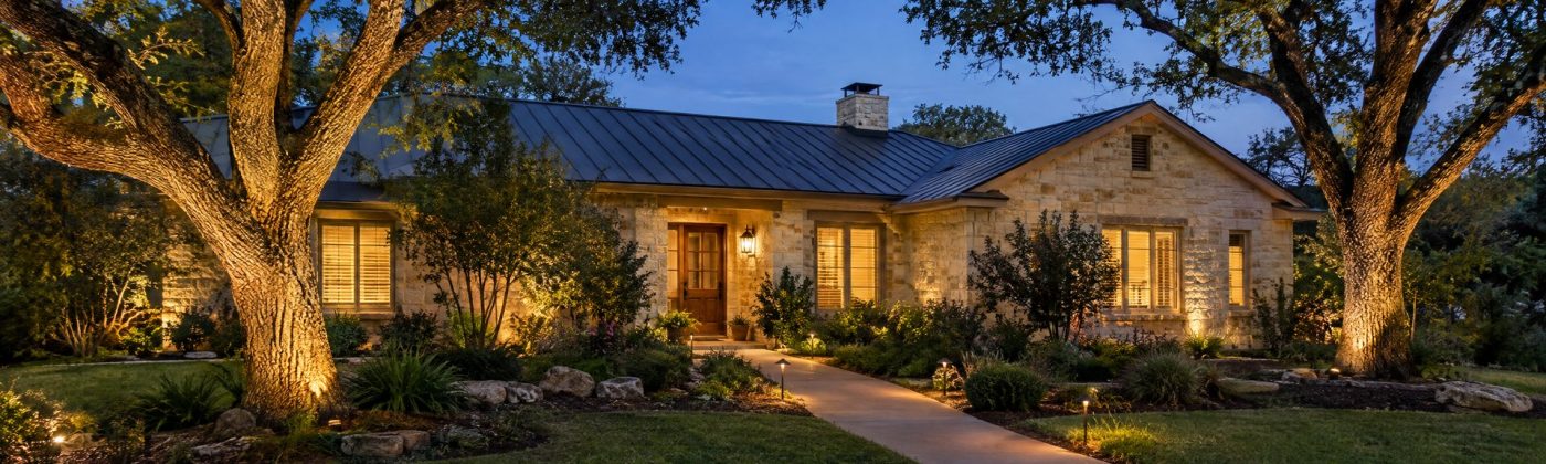 Limestone ranch house at dusk with uplit live oaks and path lights along a curved front walk
