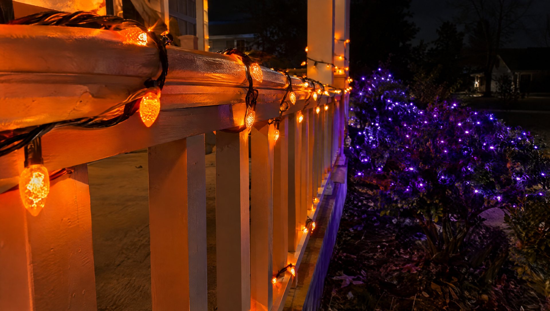 Orange-lit porch rail in front of a separate purple-lit shrub bed at night