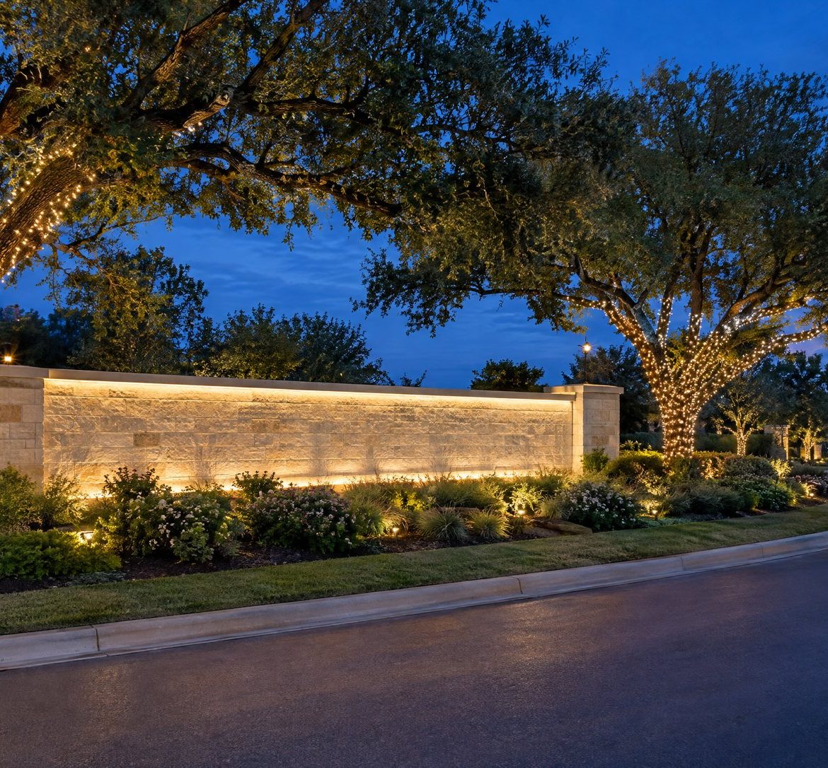Limestone subdivision entrance at blue hour with warm white lights on the wall and two wrapped live oaks