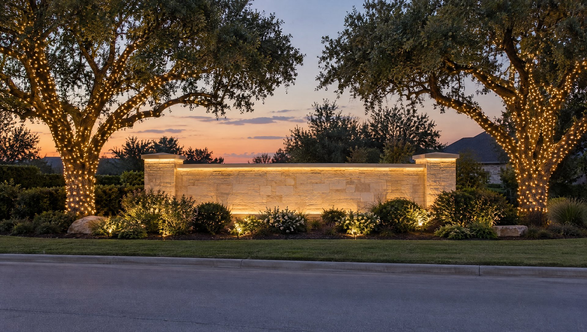 Limestone entrance monument at dusk with warm white lights along its edges and two wrapped live oaks beside it