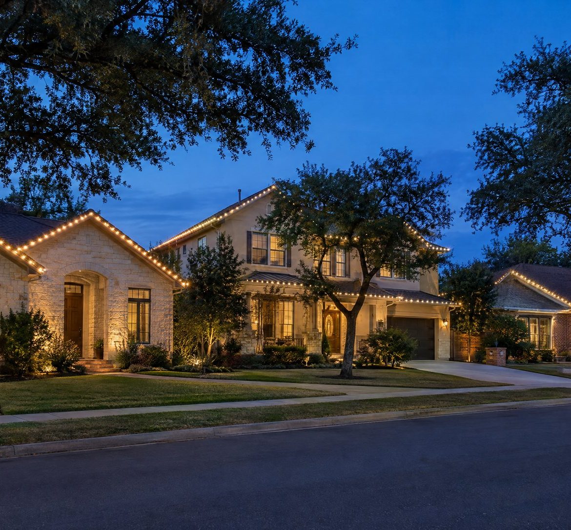 Austin street at blue hour with several homes outlined in straight warm white roofline lights.