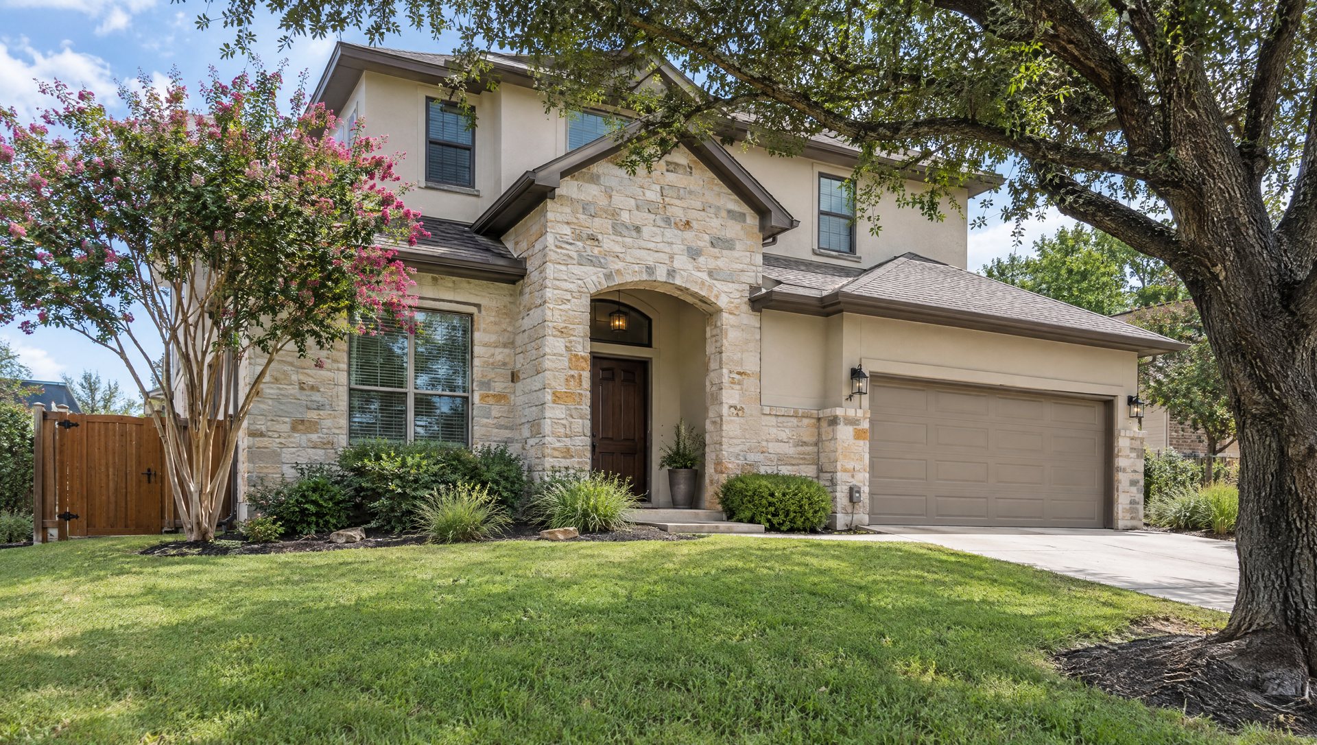Two-story stone and stucco house with a side gate, a crape myrtle and a live oak on a sloped lawn.