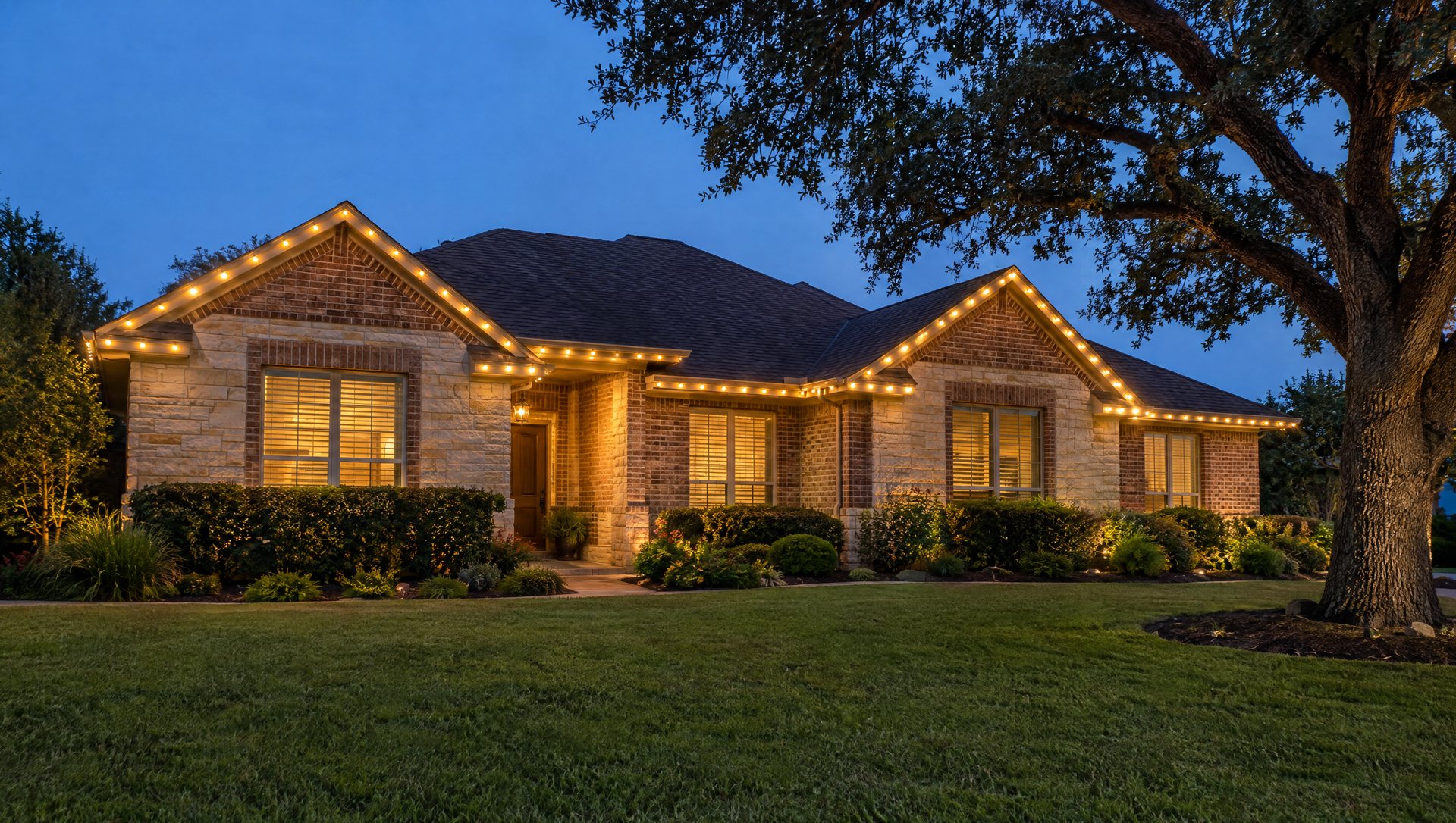 One-story brick and limestone home at blue hour with straight, evenly spaced warm white roofline bulbs.