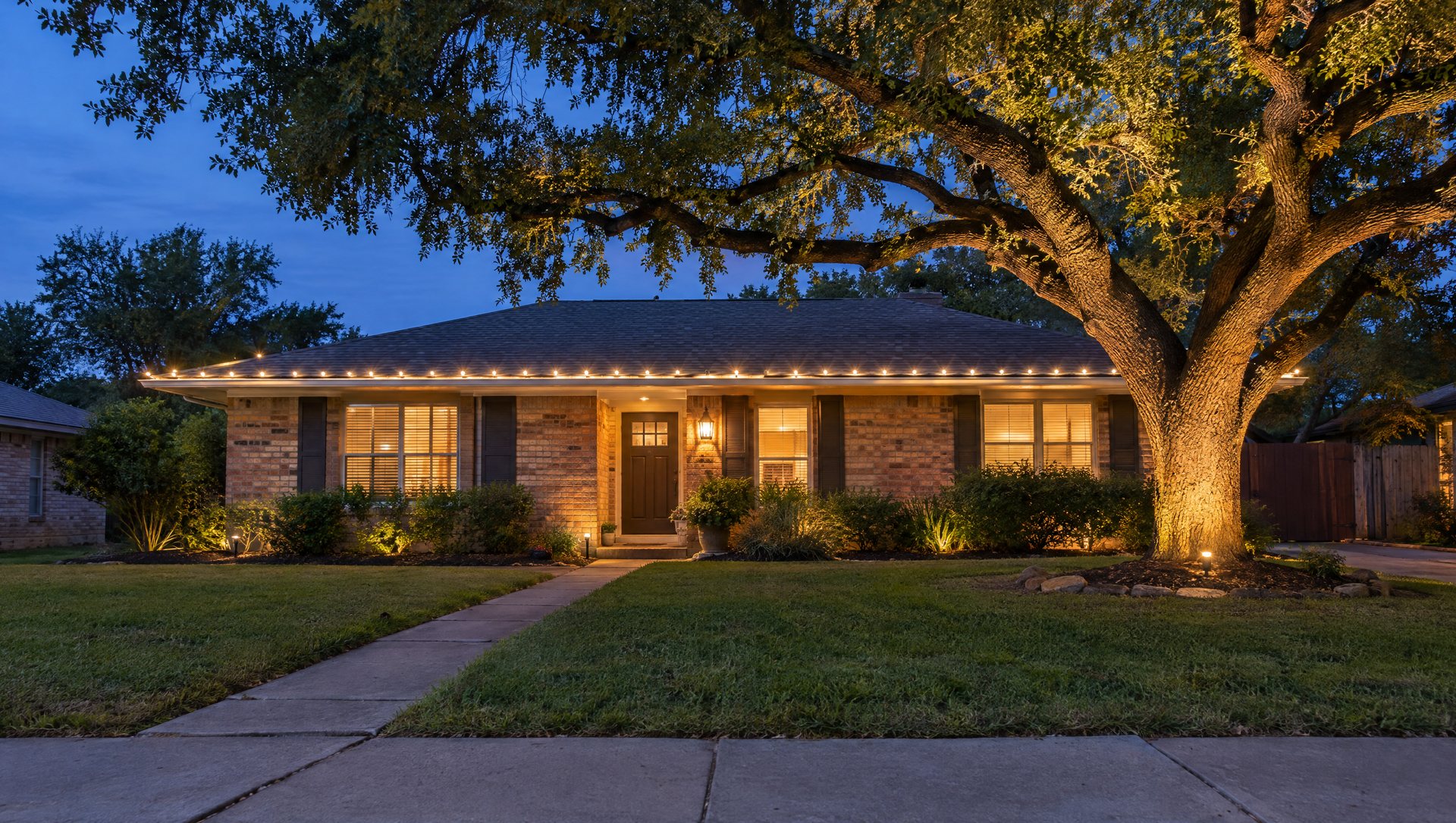 One-story brick home with warm white Christmas roofline lights and a lit tree, seen from the street at dusk
