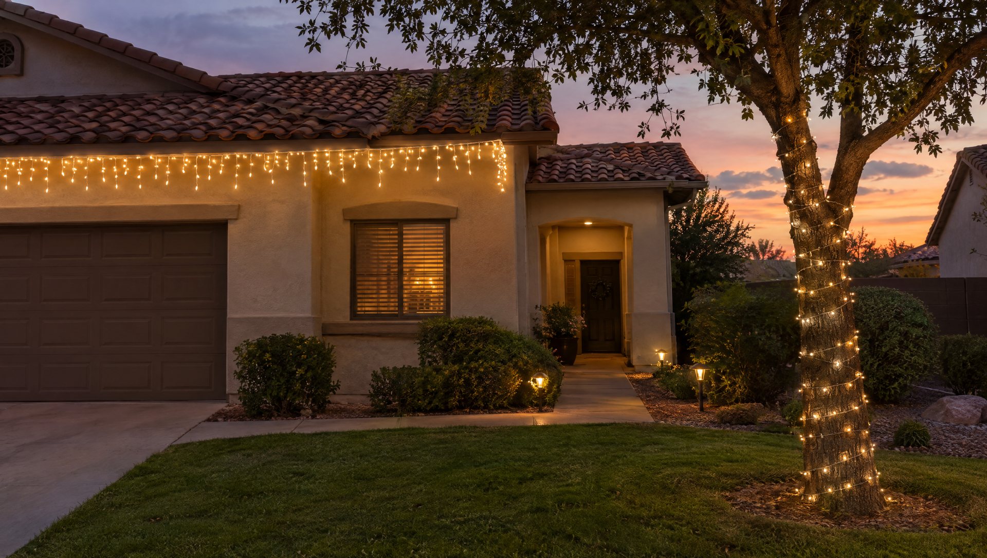 Stucco home with tile roof, icicle lights ending before the porch and a wrapped trunk