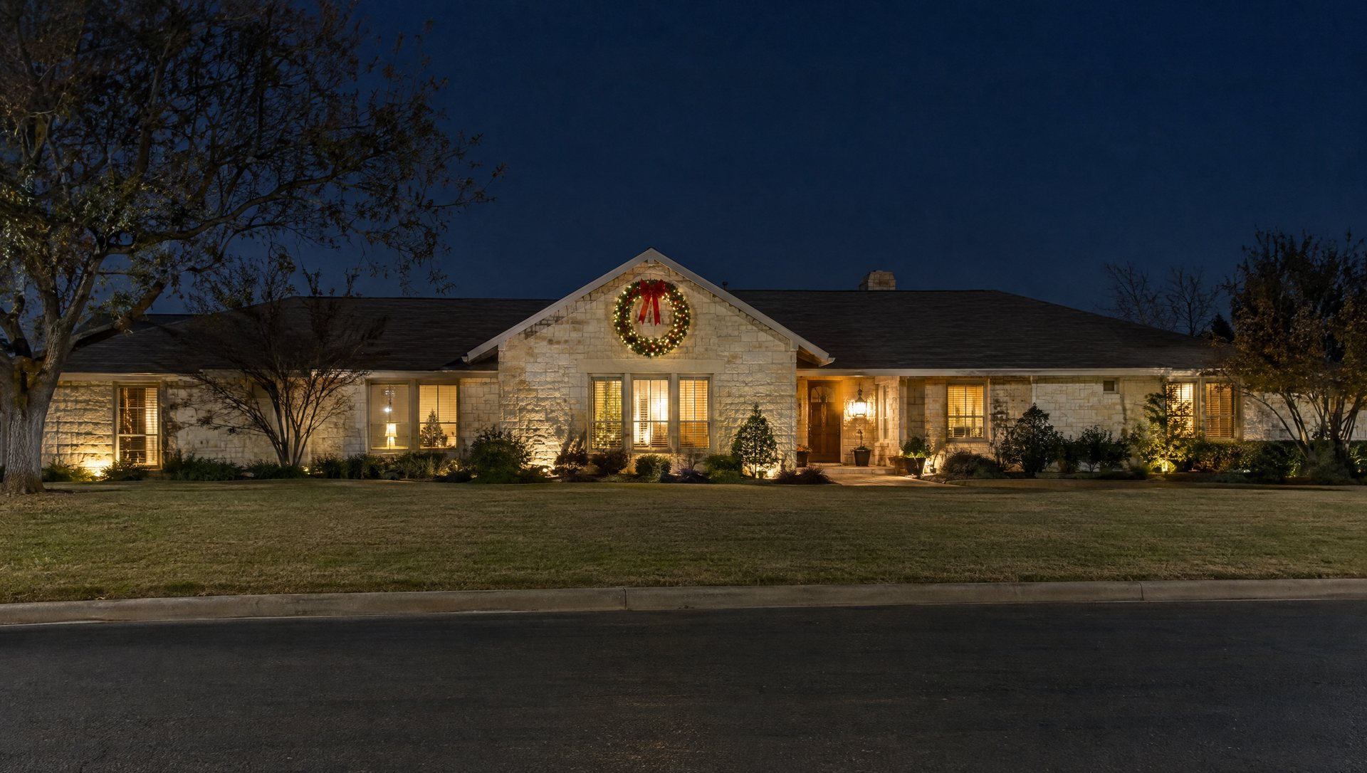 Large lit Christmas wreath hung high on the gable of a long limestone ranch house at night