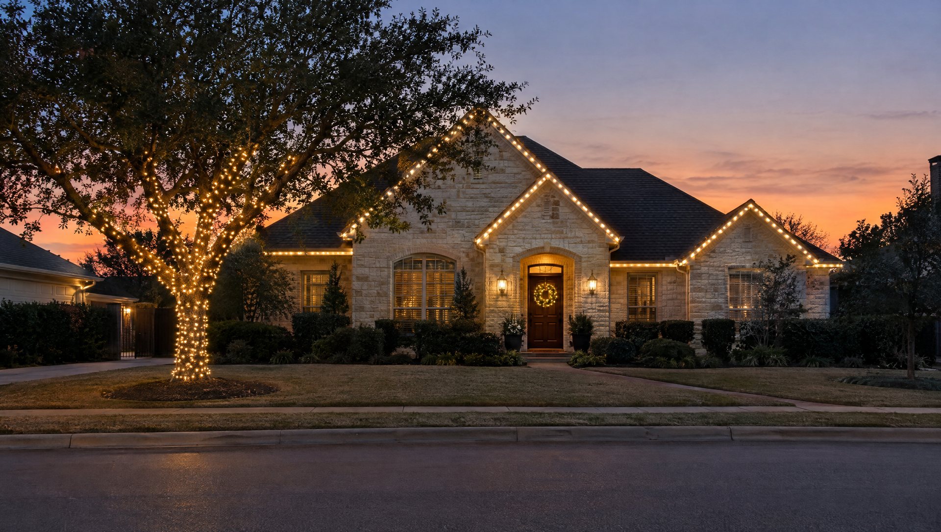 Limestone home with roofline lights, one wrapped tree and a lit wreath at dusk