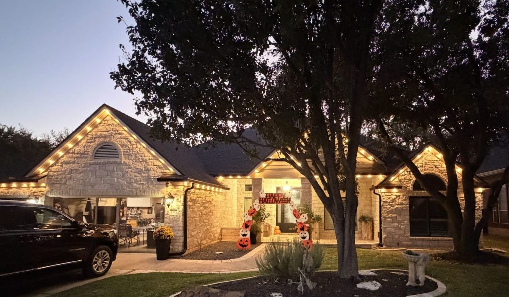 Single-story stone home at dusk with warm white bulbs along every roofline and gable