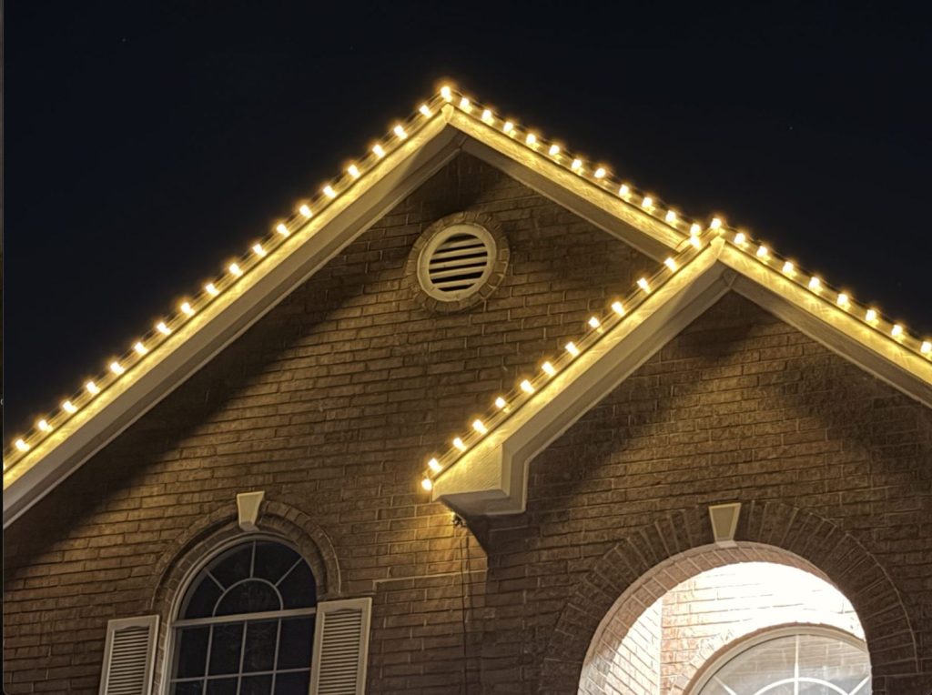 Close-up of warm white bulbs tracing a brick home's twin gables