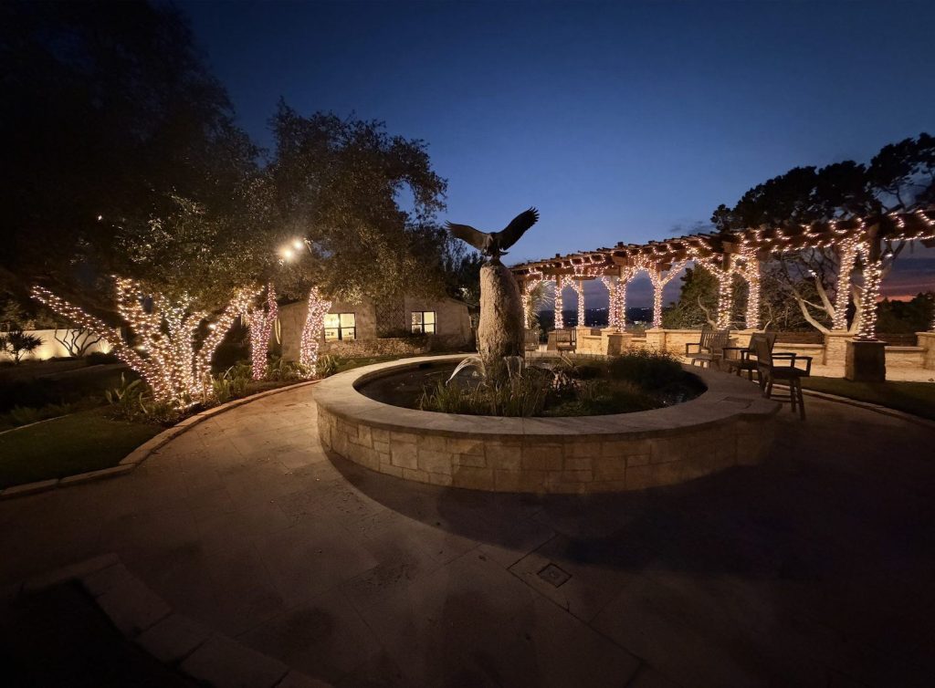 Courtyard trees and pergola wrapped in warm white lights around a fountain