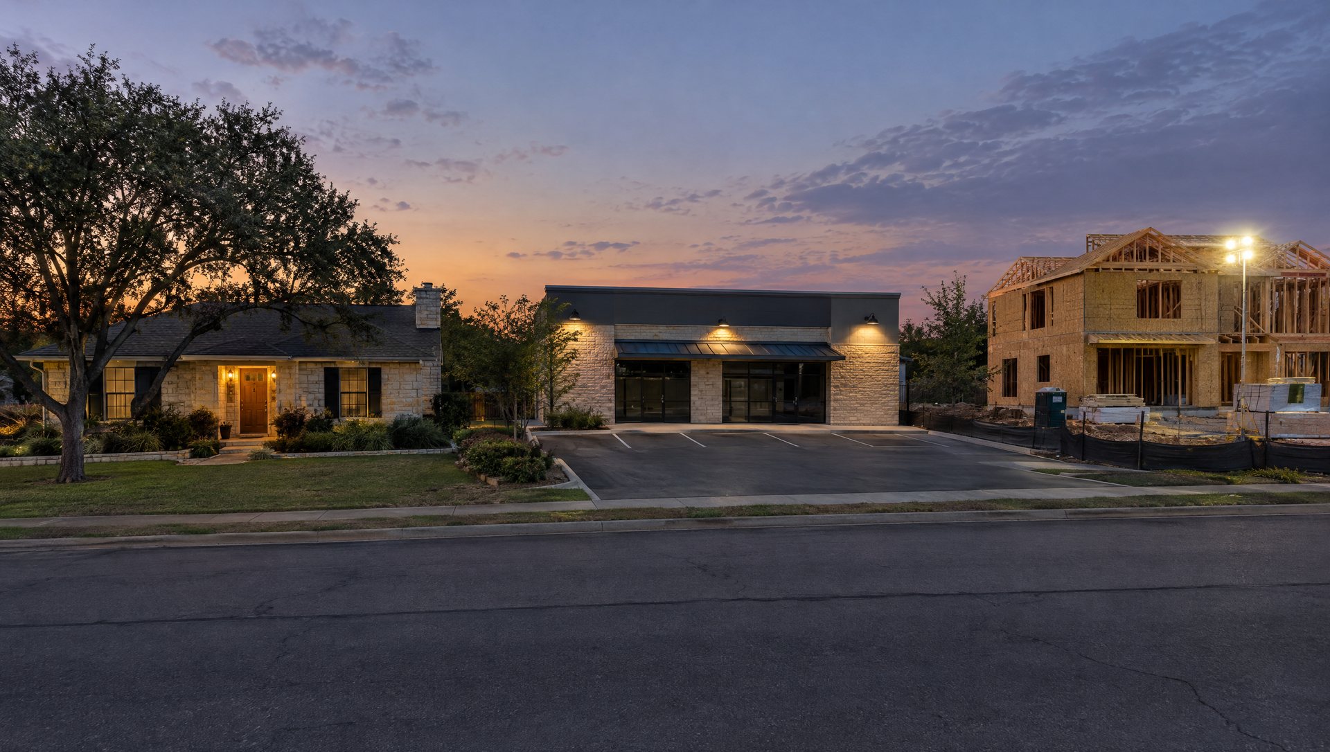 Austin street at dusk with a limestone house, a small shop with shielded parking lights and a home under construction