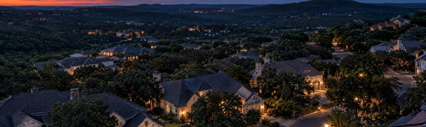 Aerial dusk view of a Hill Country neighborhood with warm, downward porch lights and dark hills under early stars