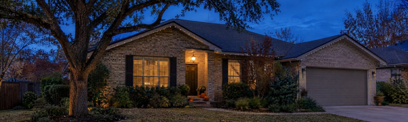 Austin brick home at blue hour with unlit Christmas bulbs along the roofline and pumpkins on the steps