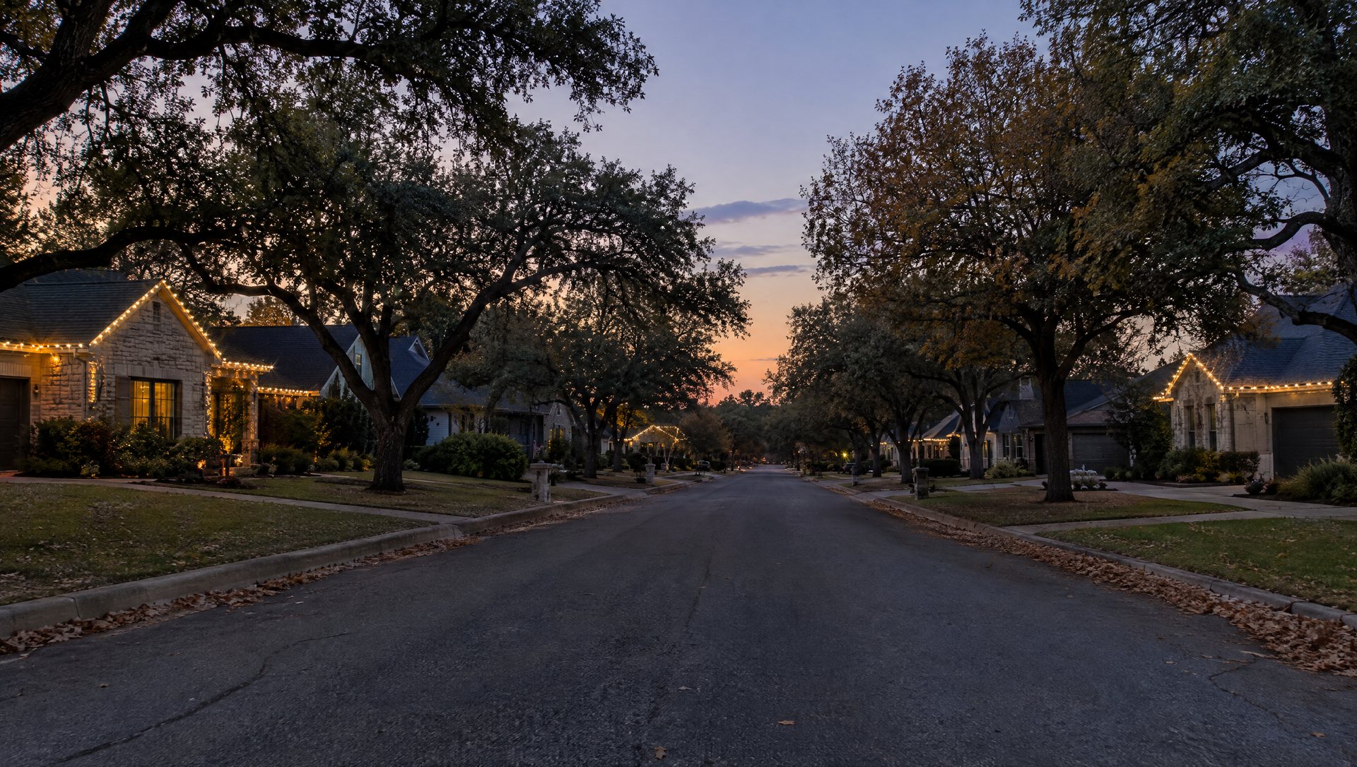 Residential street at dusk with a few homes lit for Christmas and most still dark