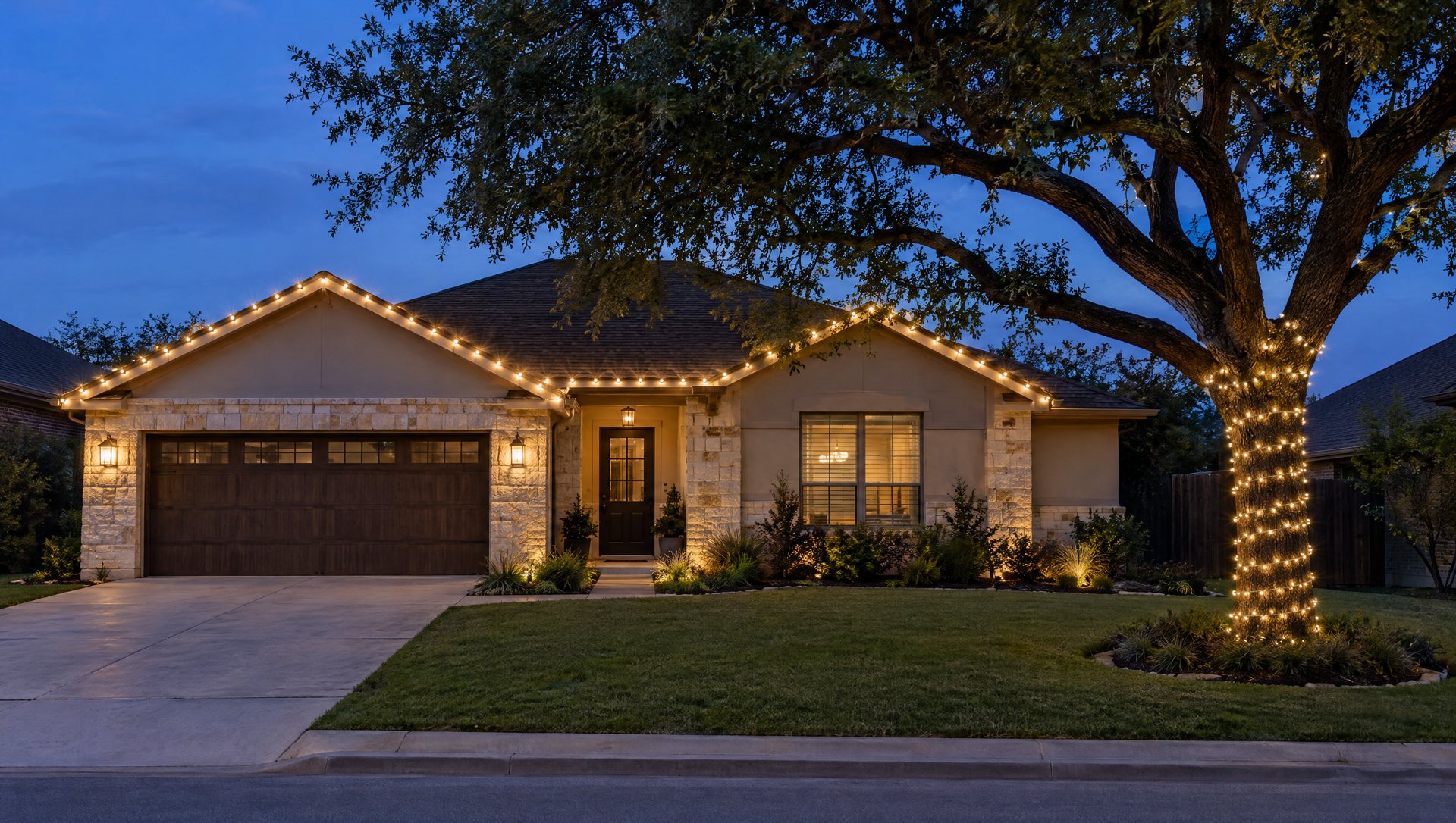 A one-story home with only its front roofline lit and one evenly wrapped live oak at blue hour.